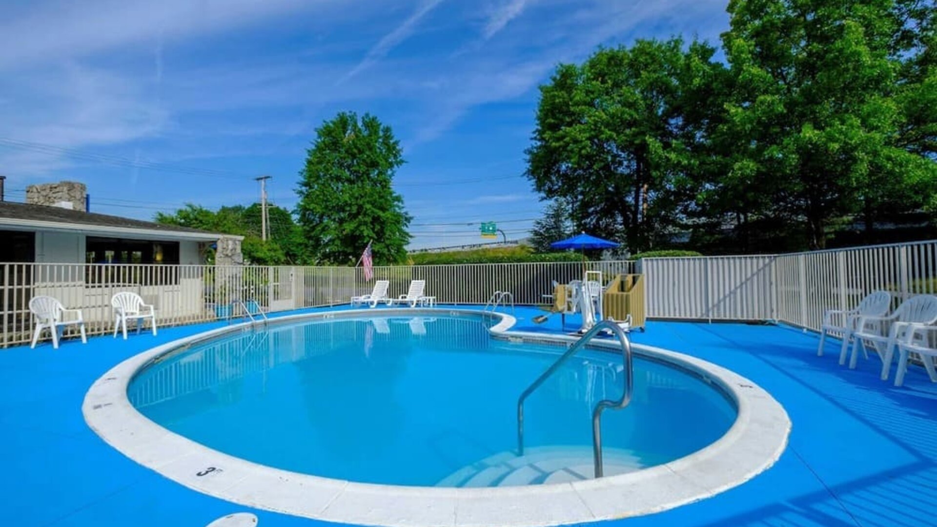 Outdoor swimming pool surrounded by a bright blue deck, white fencing, lounge chairs, and trees at American Inn Carlisle.