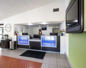 Reception desk with American Inn Carlisle signage, computers, a wall-mounted television, and a tiled lobby floor.