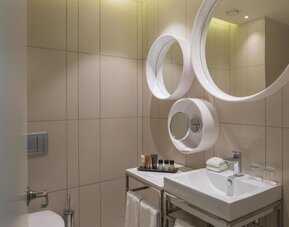 Contemporary bathroom with beige tiled walls, a white vanity sink, circular mirrors, toiletries, and neatly folded towels at Prima Millennium.