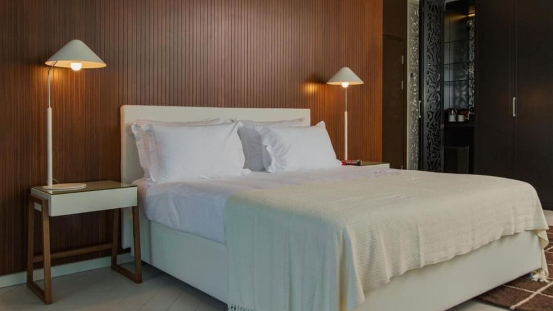 King bedroom featuring white bedding, a wood-panelled feature wall, bedside tables, matching lamps, and dark built-in wardrobes at Prima Millennium.