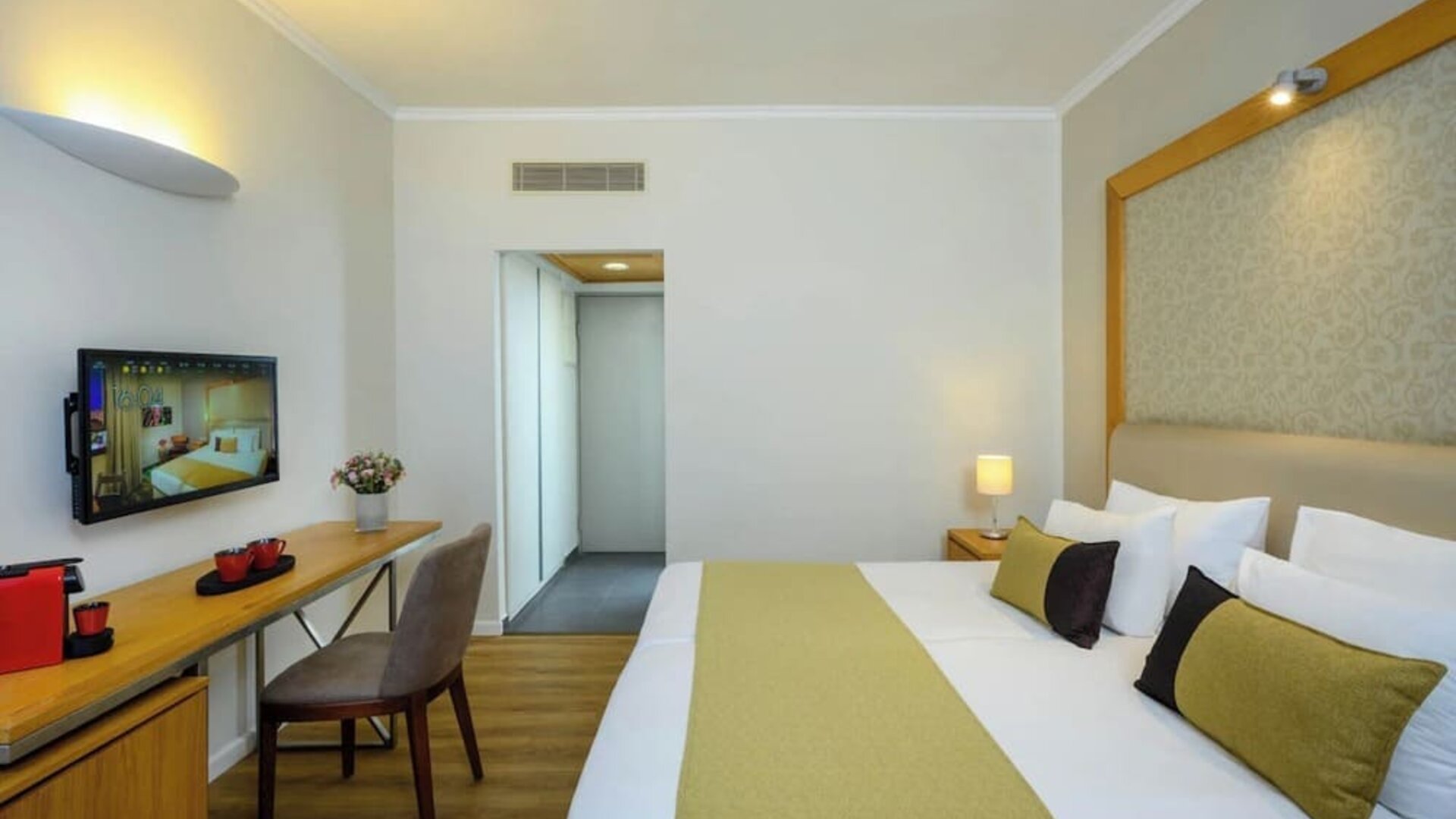 Guest bedroom with a king-size bed, white bedding, a gold bed runner, bedside tables, a wall-mounted television, and a desk with a chair at Prima Park Hotel.