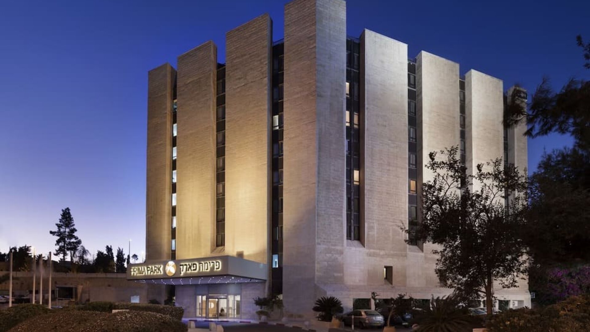Exterior view of Prima Park Hotel at dusk, showing the multi-storey building illuminated against the evening sky, landscaped grounds, and main entrance.