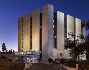Exterior view of Prima Park Hotel at dusk, showing the multi-storey building illuminated against the evening sky, landscaped grounds, and main entrance.