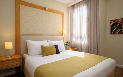 Guest room featuring a king-size bed with white bedding, a gold bed runner, decorative accent cushions, wooden bedside tables, and a large upholstered headboard at Prima Park Hotel.