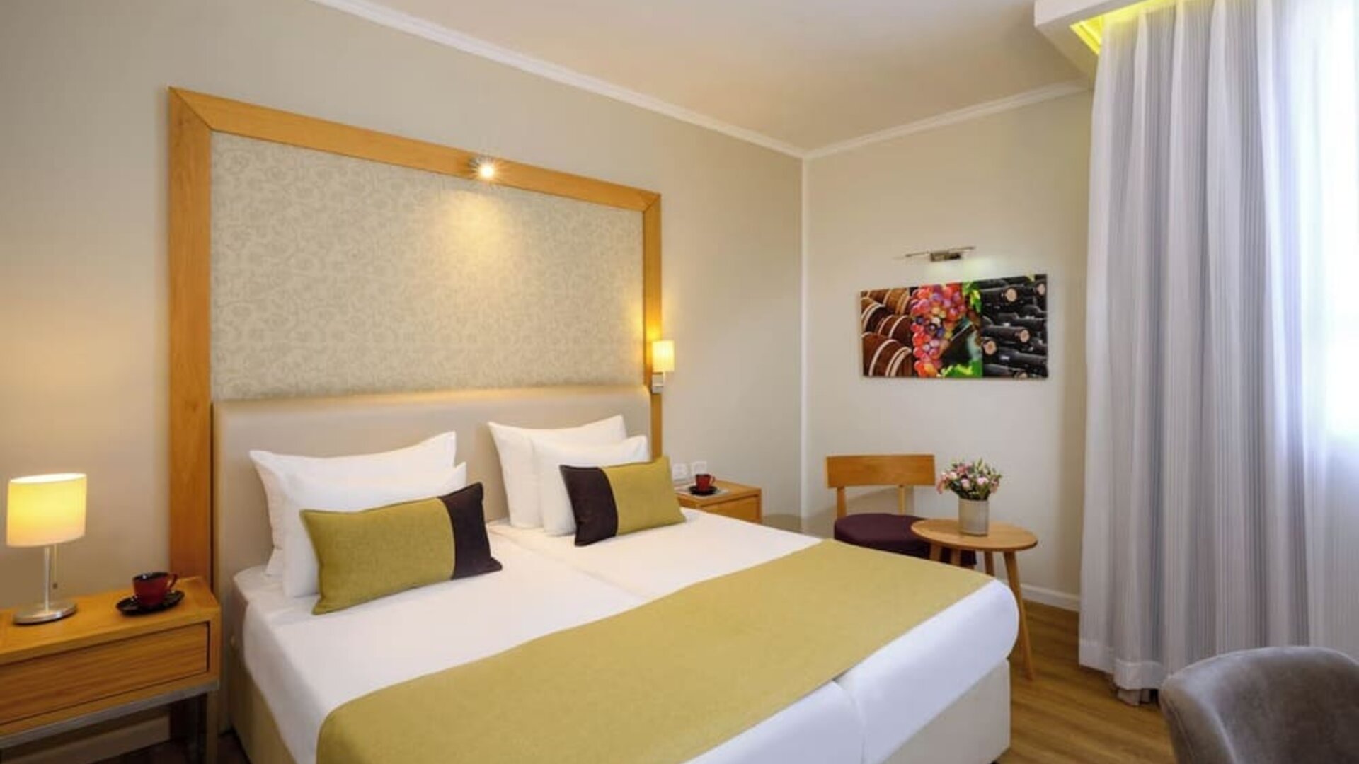 Guest room with a king-size bed, white bedding, gold and dark accent cushions, a wooden headboard frame, and a small seating area beside the window at Prima Park Hotel.