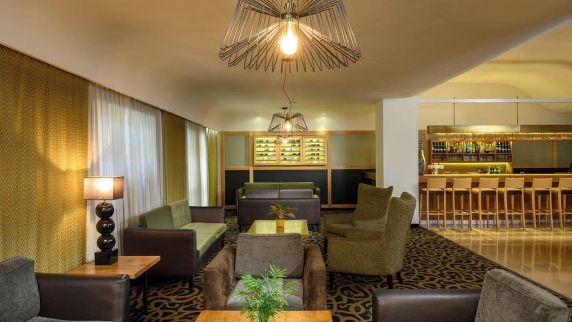 Hotel lounge with upholstered armchairs and sofas, wooden coffee tables, patterned carpeting, decorative pendant lighting, and an adjoining bar at Prima Park Hotel.