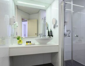 Bright hotel bathroom featuring a white vanity with a vessel sink, large mirror, glass-enclosed shower, wall-mounted hairdryer, and toiletries at Prima Park Hotel.