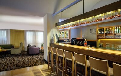 Hotel bar with a long wooden counter, bar stools, illuminated bottle shelves, glassware, and an adjoining lounge seating area at Prima Park Hotel.