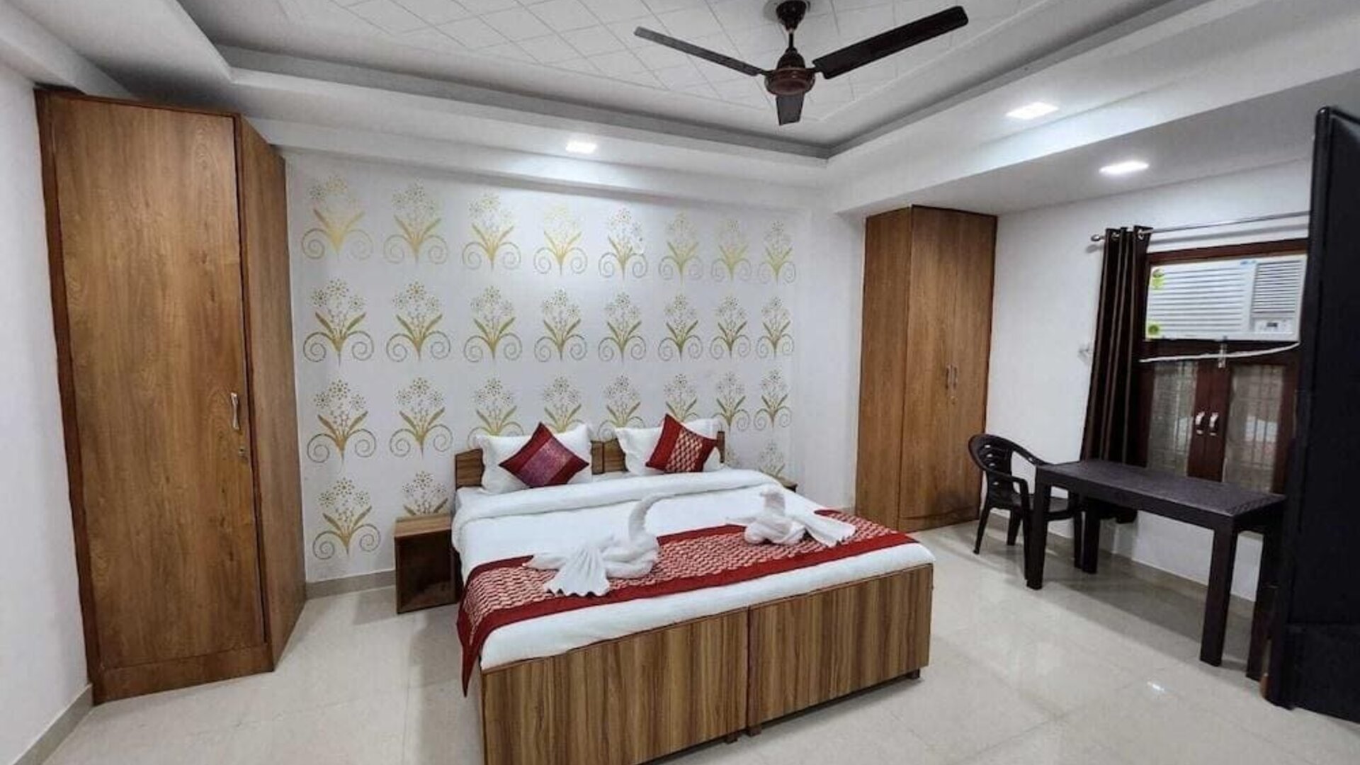 Guest bedroom with a wooden double bed, white bedding, red decorative cushions and bed runner, patterned feature wall, wardrobes, ceiling fan, and a small table with chairs beside the window at Hotel Urban Abode.