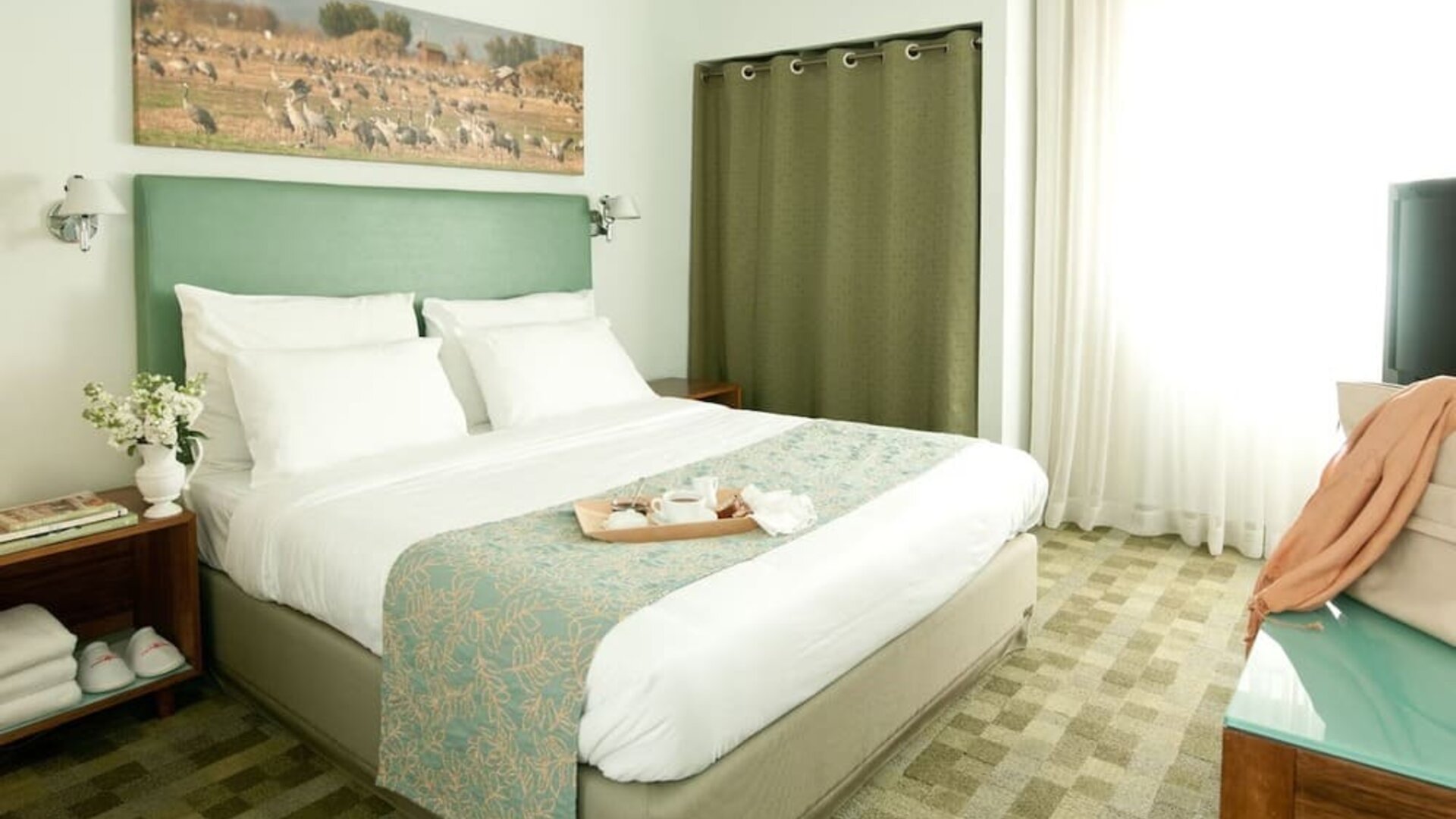 Guest bedroom featuring a king bed, sage-green headboard, patterned runner and bright windows at Prima Galil Tiberias.