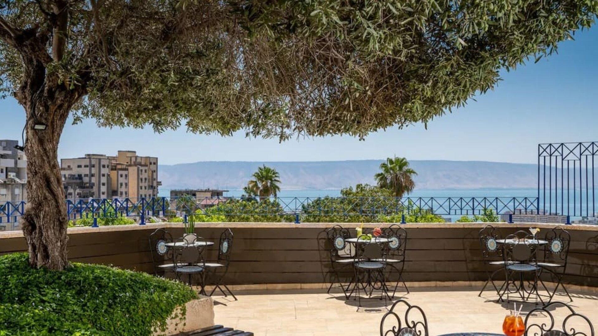 Outdoor terrace with café seating, mature trees and views of the Sea of Galilee at Prima Galil Tiberias.