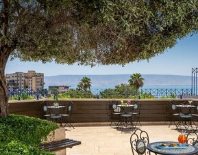 Outdoor terrace with café seating, mature trees and views of the Sea of Galilee at Prima Galil Tiberias.