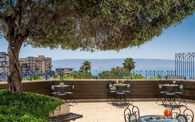 Outdoor terrace with café seating, mature trees and views of the Sea of Galilee at Prima Galil Tiberias.