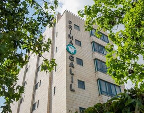Exterior view of the hotel building surrounded by mature green trees at Prima Galil Tiberias.