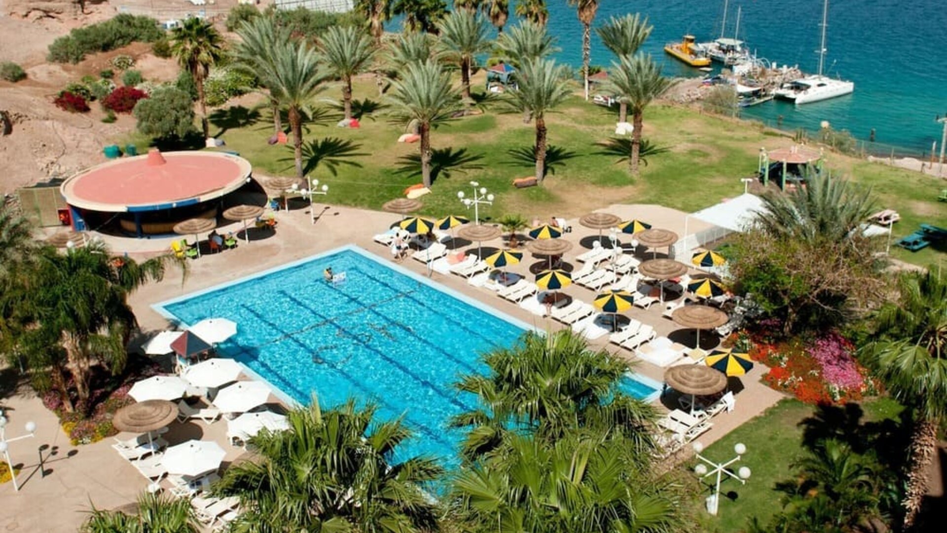Aerial view of the outdoor swimming pool surrounded by palm trees, sun loungers and landscaped gardens beside the Red Sea at Prima Music Eilat