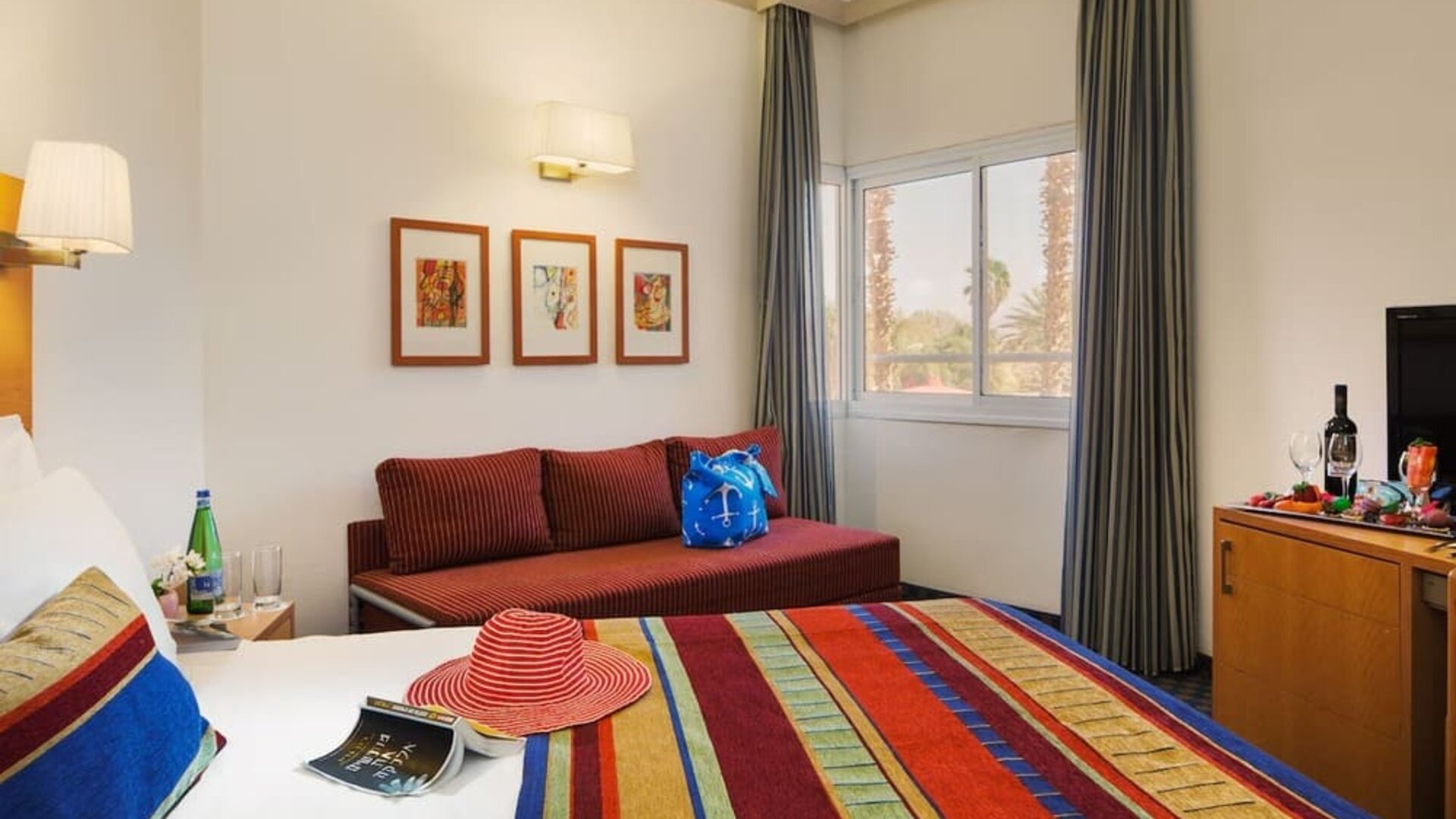 Guest bedroom with a colourful striped bedspread, red sofa, framed artwork and a flat-screen television at Prima Music Eilat.