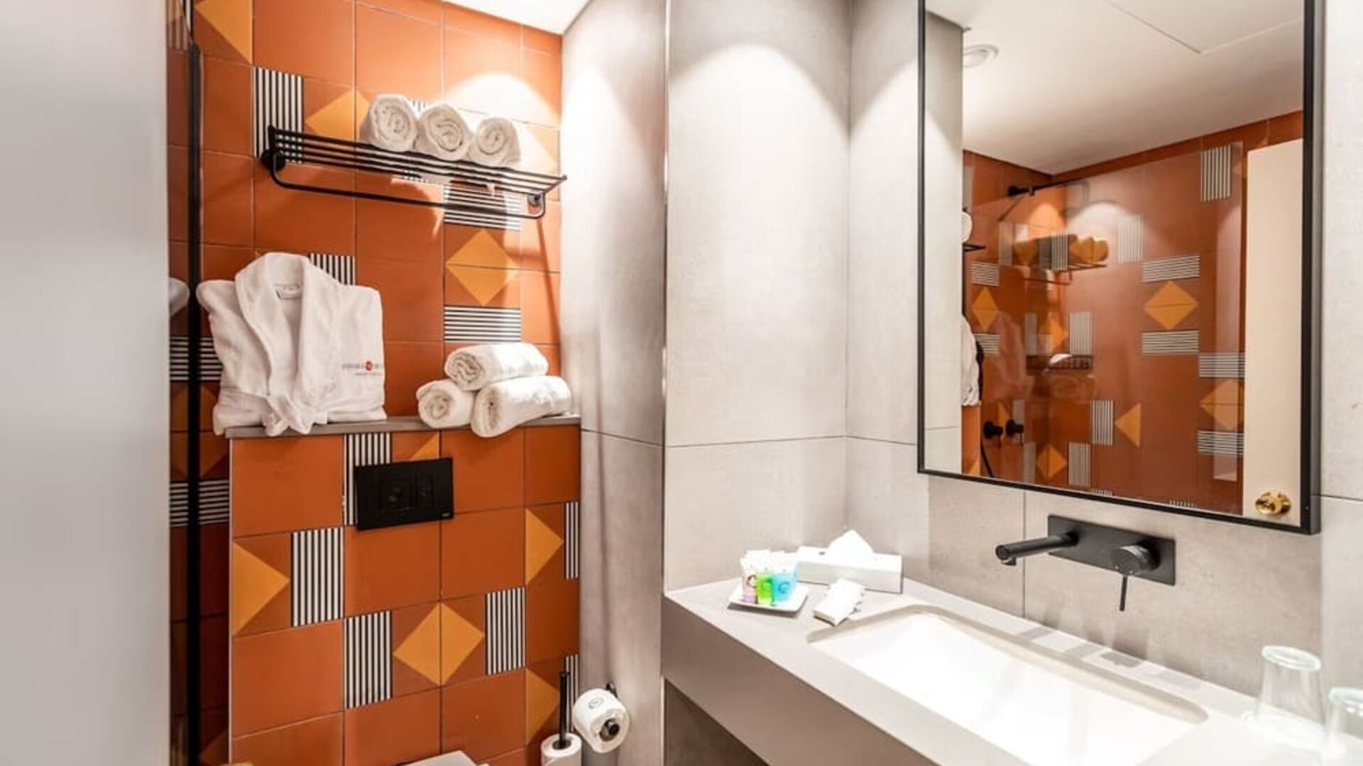 Bathroom with orange geometric accent tiles, a wall-mounted towel rack, bathrobes and a wide vanity mirror at Prima Music Eilat.