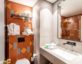 Bathroom with orange geometric accent tiles, a wall-mounted towel rack, bathrobes and a wide vanity mirror at Prima Music Eilat.