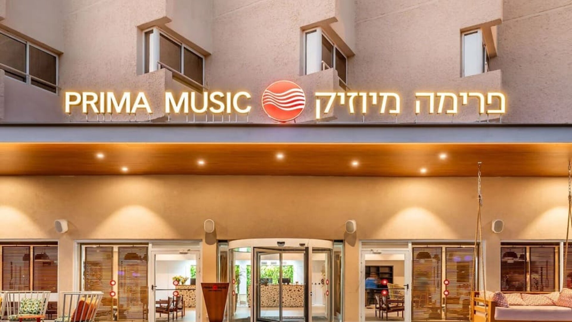 Exterior entrance to Prima Music Eilat, featuring illuminated hotel signage and glass entrance doors.