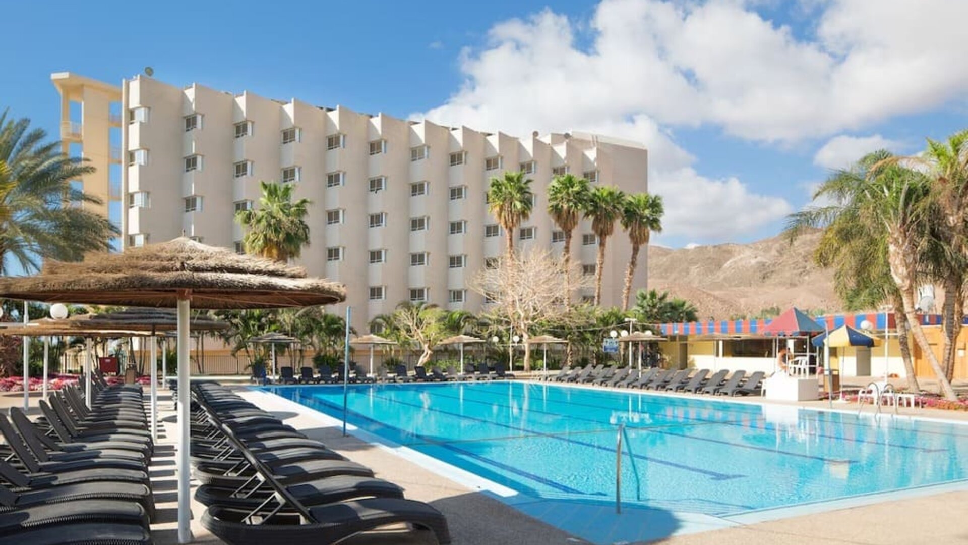 Outdoor swimming pool with rows of sun loungers, parasols and views of the hotel building and surrounding desert hills at Prima Music Eilat.