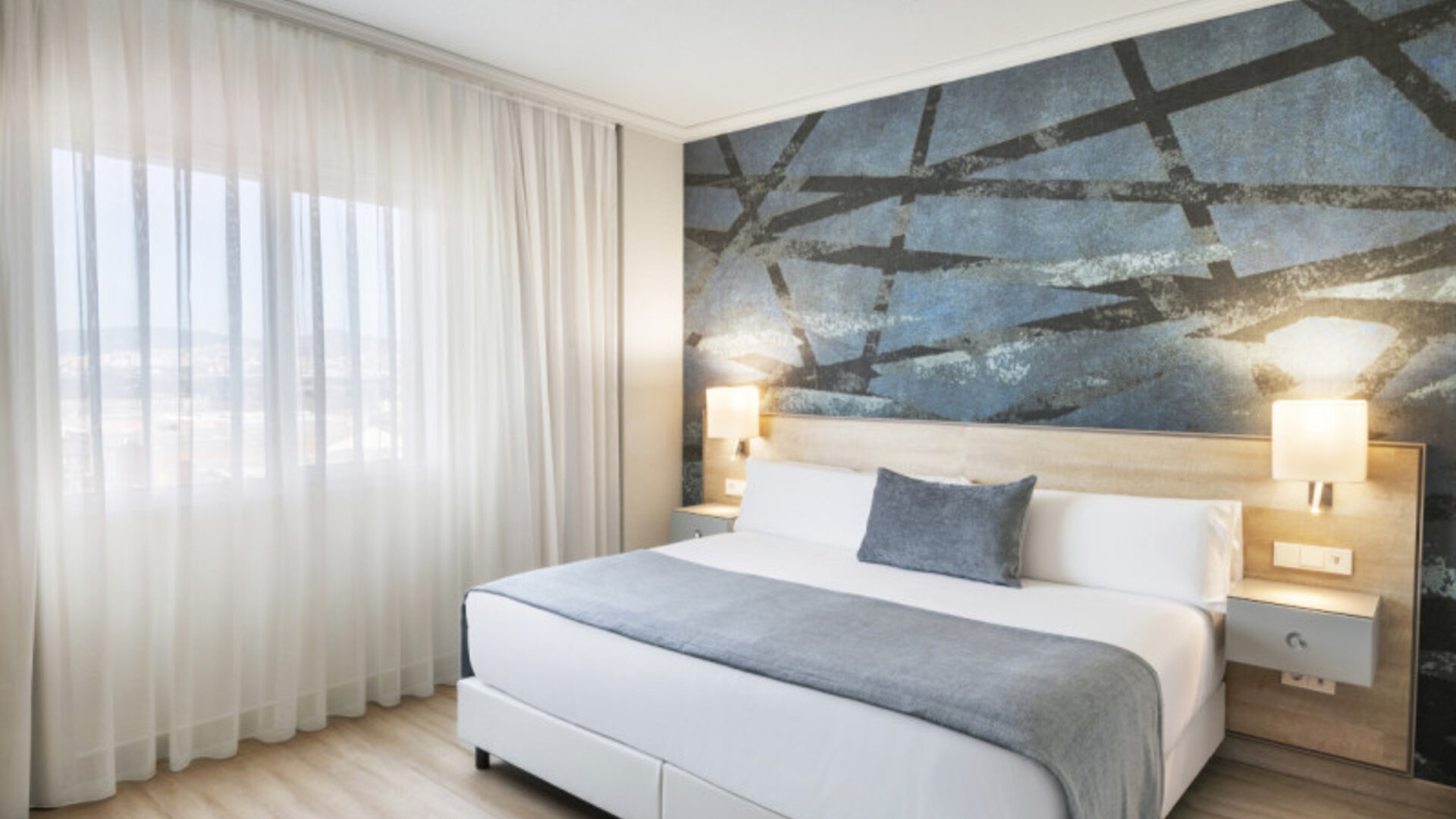 King-size bed with white bedding, grey accents, sheer curtains and a dark abstract feature wall at Hotel Sallés Ciutat del Prat.