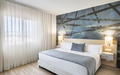 King-size bed with white bedding, grey accents, sheer curtains and a dark abstract feature wall at Hotel Sallés Ciutat del Prat.