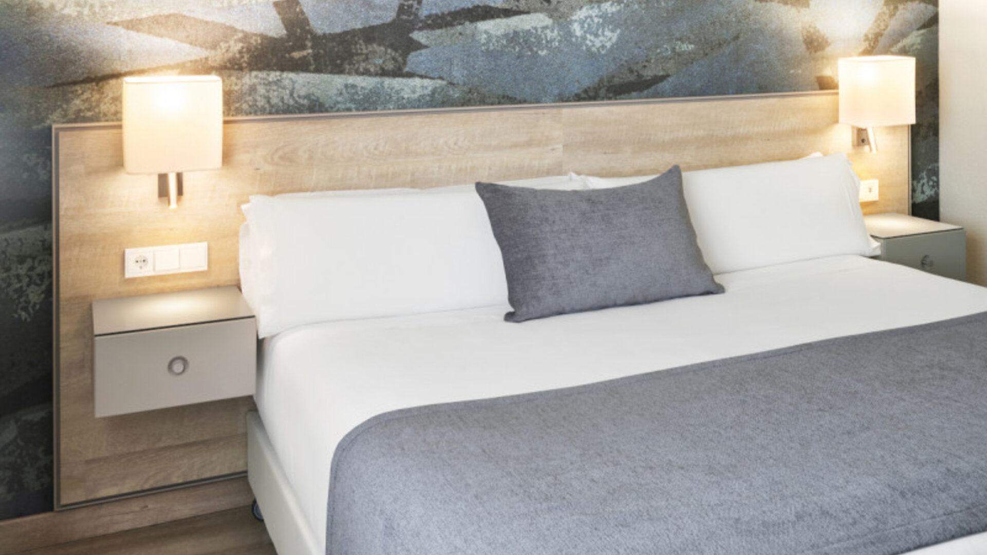 King-size bed with white linens, grey throw and cushion, illuminated bedside lamps and an abstract feature wall at Hotel Sallés Ciutat del Prat.