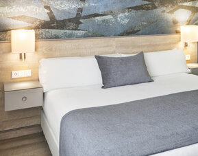 King-size bed with white linens, grey throw and cushion, illuminated bedside lamps and an abstract feature wall at Hotel Sallés Ciutat del Prat.