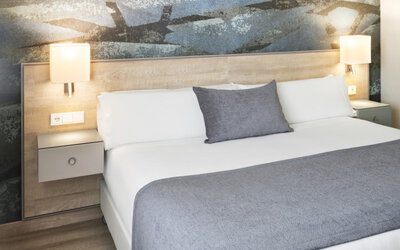 King-size bed with white linens, grey throw and cushion, illuminated bedside lamps and an abstract feature wall at Hotel Sallés Ciutat del Prat.