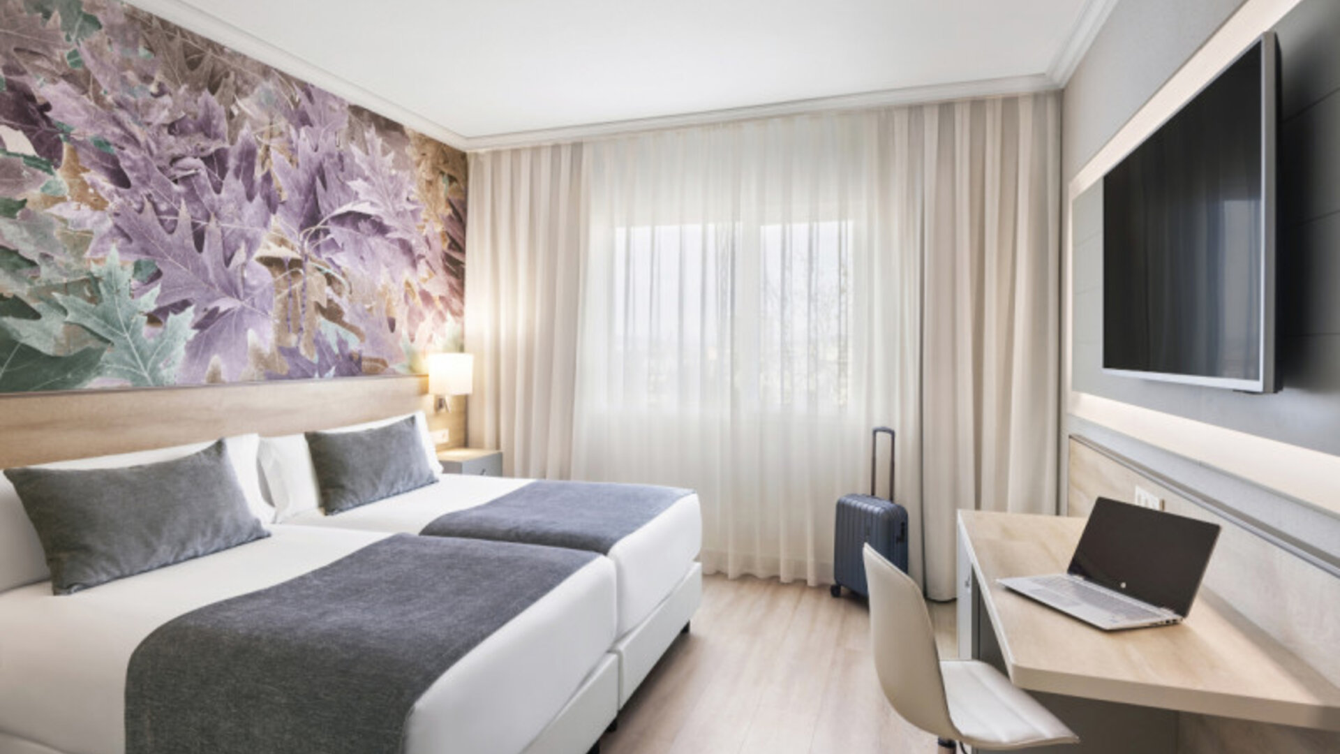 Guest room with twin beds, grey bedding, a work desk, open suitcase and decorative botanical wall art at Hotel Sallés Ciutat del Prat.