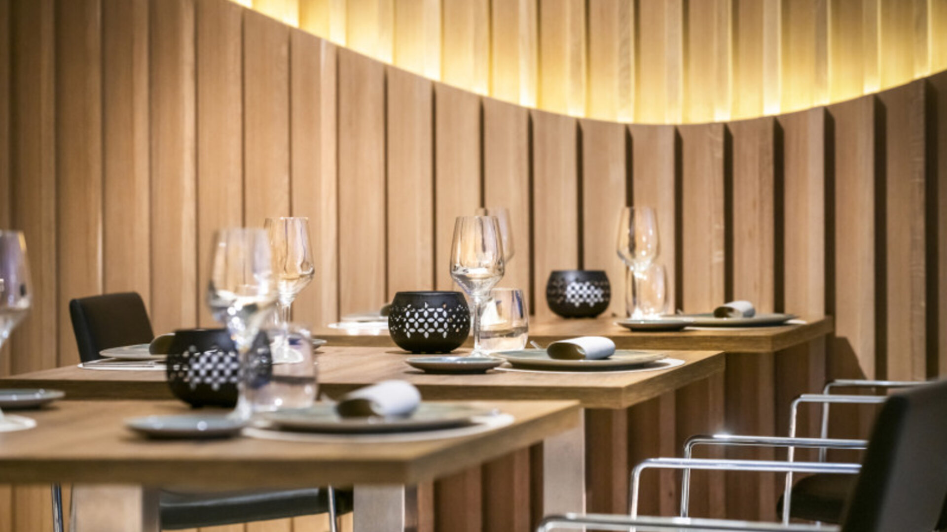 Restaurant tables set with plates, glasses and black chairs beside a softly illuminated wooden feature wall at Hotel Sallés Ciutat del Prat.