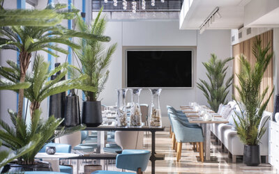Bright dining lounge with blue and cream chairs, round tables, indoor palms and floor-to-ceiling windows at Hotel Sallés Ciutat del Prat.