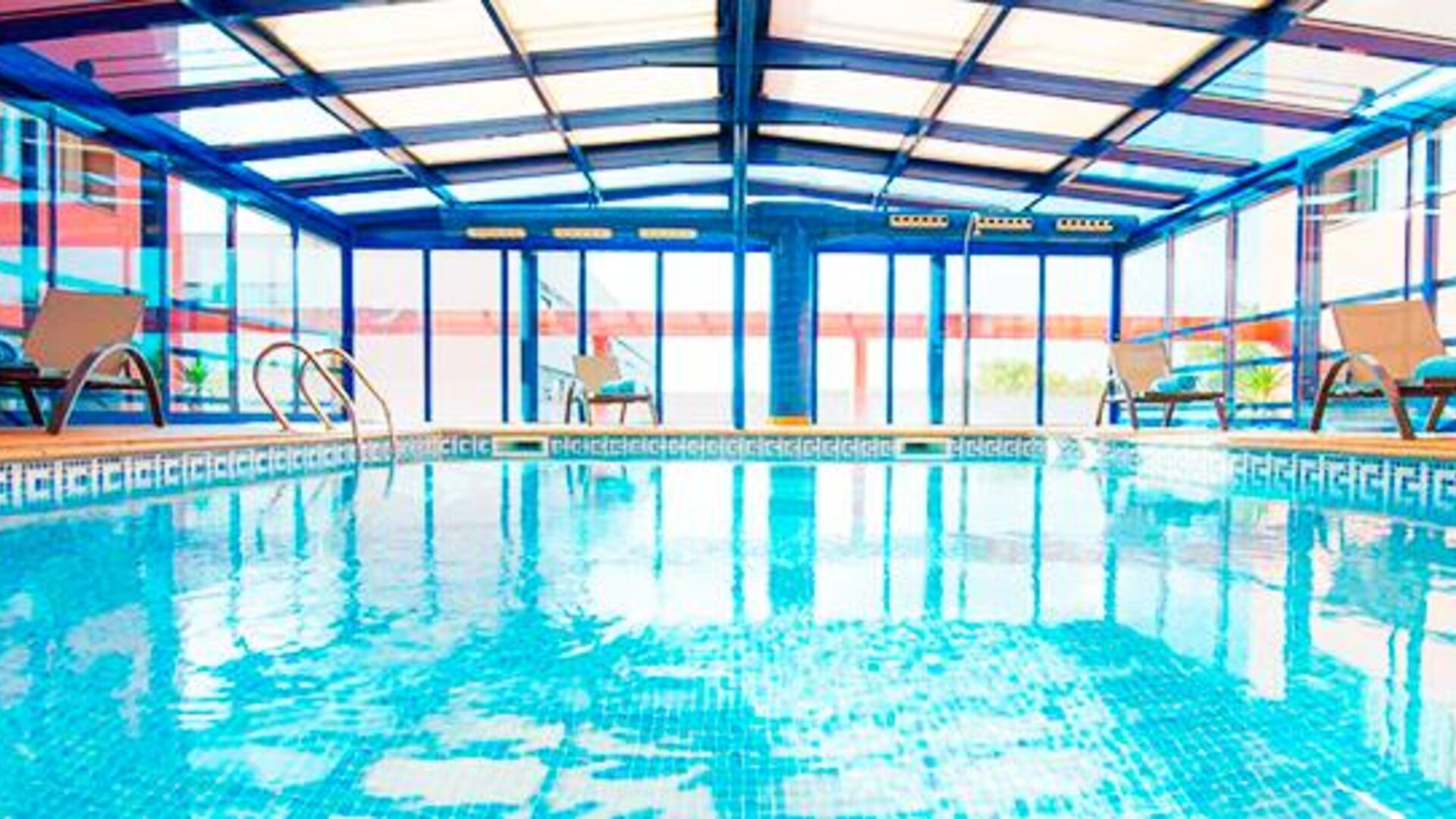 Indoor swimming pool beneath a glass roof, surrounded by tiled flooring and large windows at Hotel Sallés Ciutat del Prat.