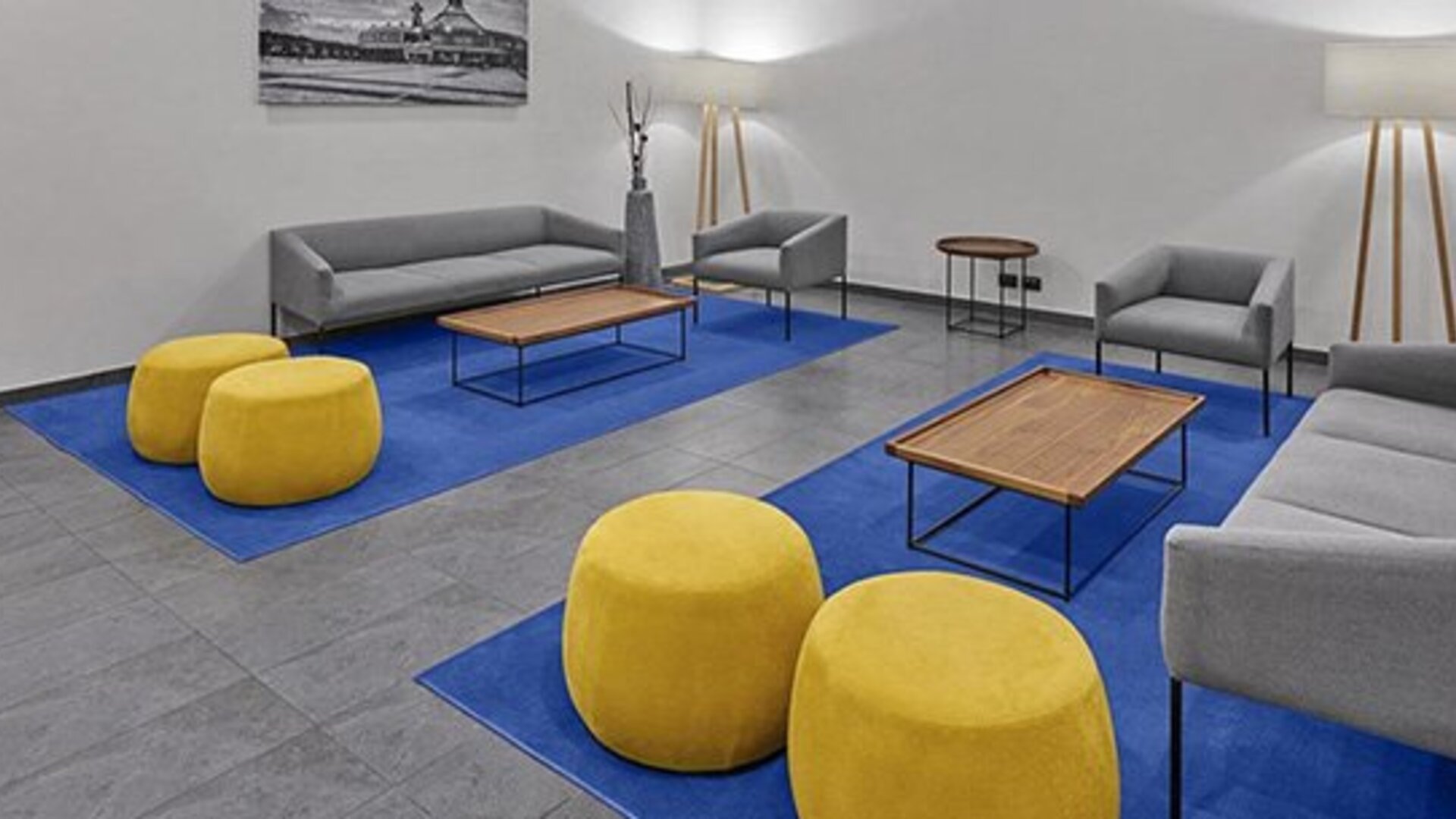 Lounge with grey sofas, yellow round ottomans, wooden coffee tables and blue carpeting at City Express by Marriott CDMX La Villa.