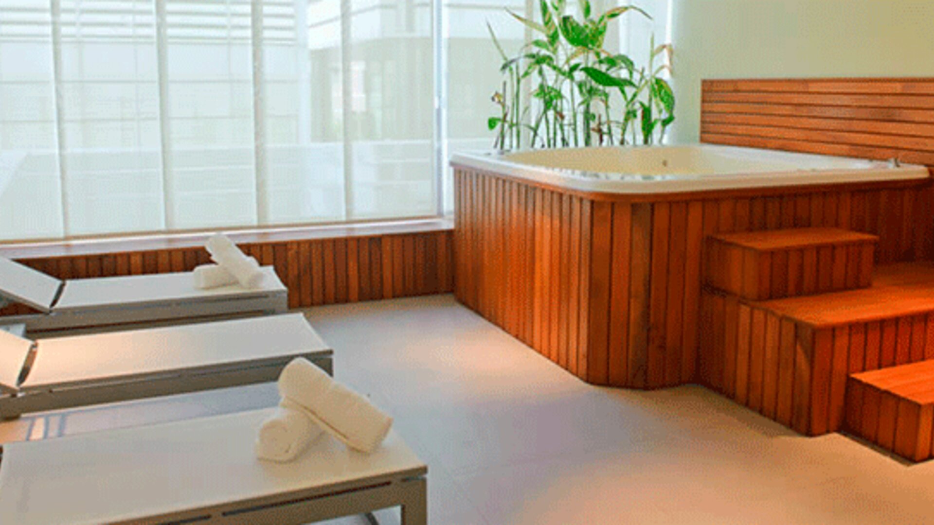 Spa relaxation area with white loungers, rolled towels, wooden hot tub and large windows at Design Suites Buenos Aires.