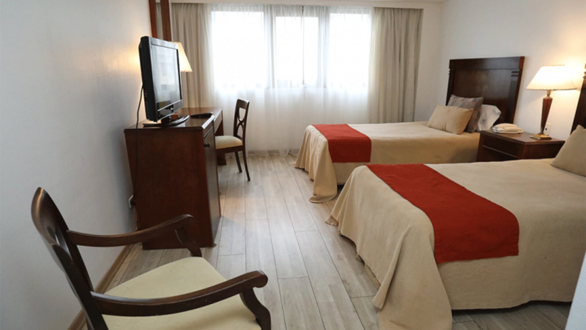 Twin guest room with two beds, orange runners, wooden furniture, television and large curtained windows at HR Esmeralda Luxor.