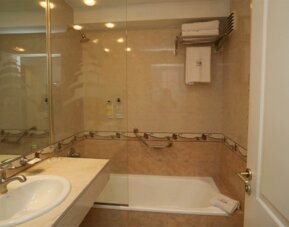 Bathroom with beige tiled walls, glass shower screen, bathtub, white vanity sink and grab rails at HR Esmeralda Luxor.