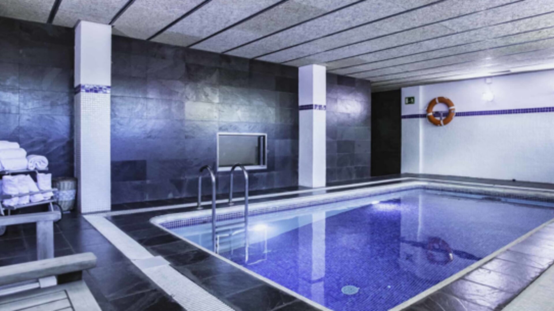 Indoor swimming pool with blue tiled water, dark stone walls, metal handrails and a towel rack at Hotel Best Western PLUS Alfa Aeropuerto.