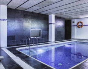 Indoor swimming pool with blue tiled water, dark stone walls, metal handrails and a towel rack at Hotel Best Western PLUS Alfa Aeropuerto.