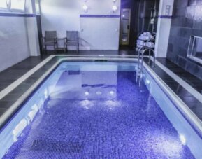 Indoor swimming pool with blue mosaic tiles, dark stone flooring, white walls and a metal handrail at Hotel Best Western PLUS Alfa Aeropuerto.