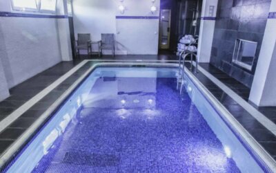 Indoor swimming pool with blue mosaic tiles, dark stone flooring, white walls and a metal handrail at Hotel Best Western PLUS Alfa Aeropuerto.