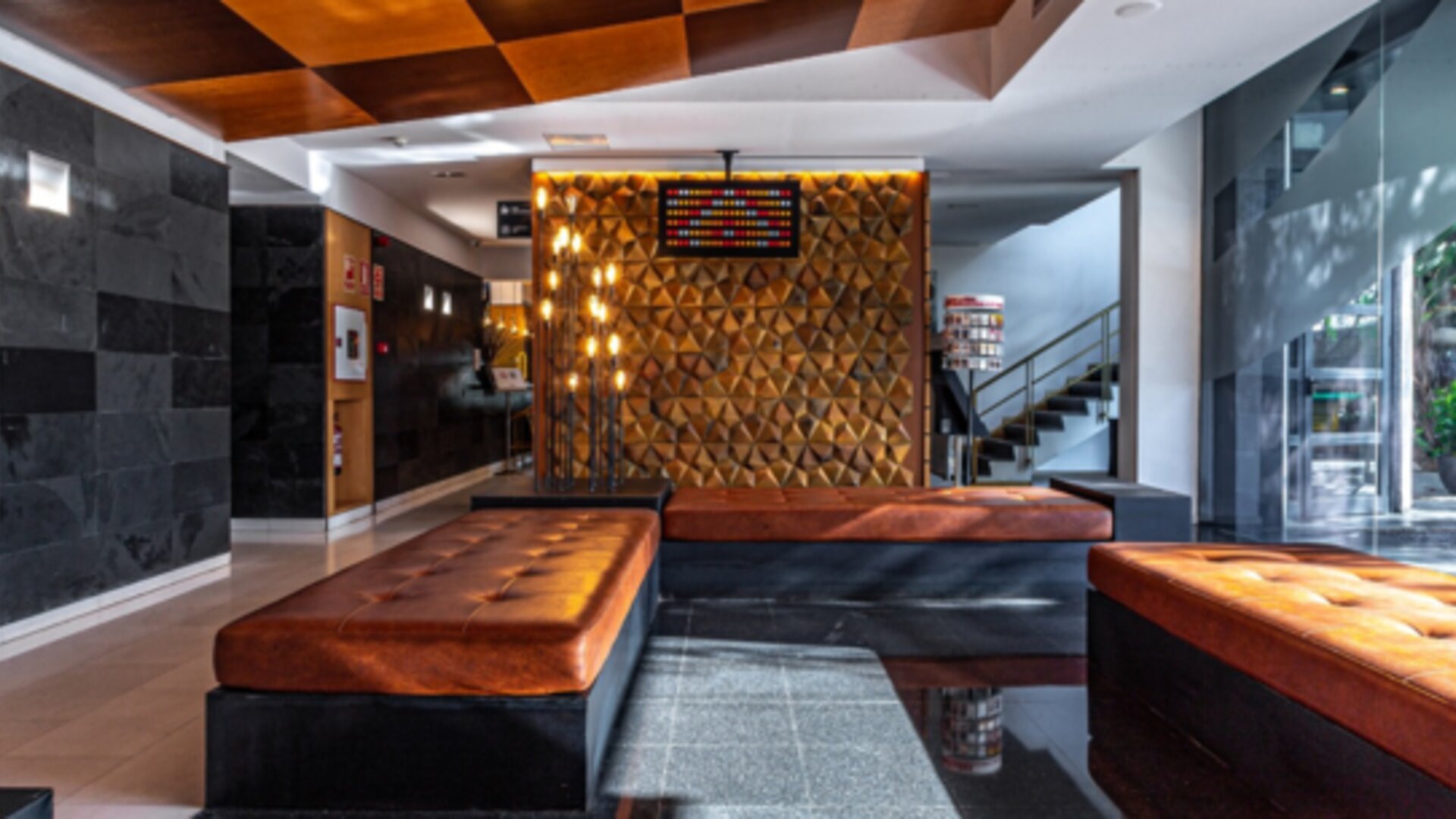 Hotel lobby with brown leather seating, geometric feature wall, staircase and large windows overlooking greenery at Hotel Best Western PLUS Alfa Aeropuerto.