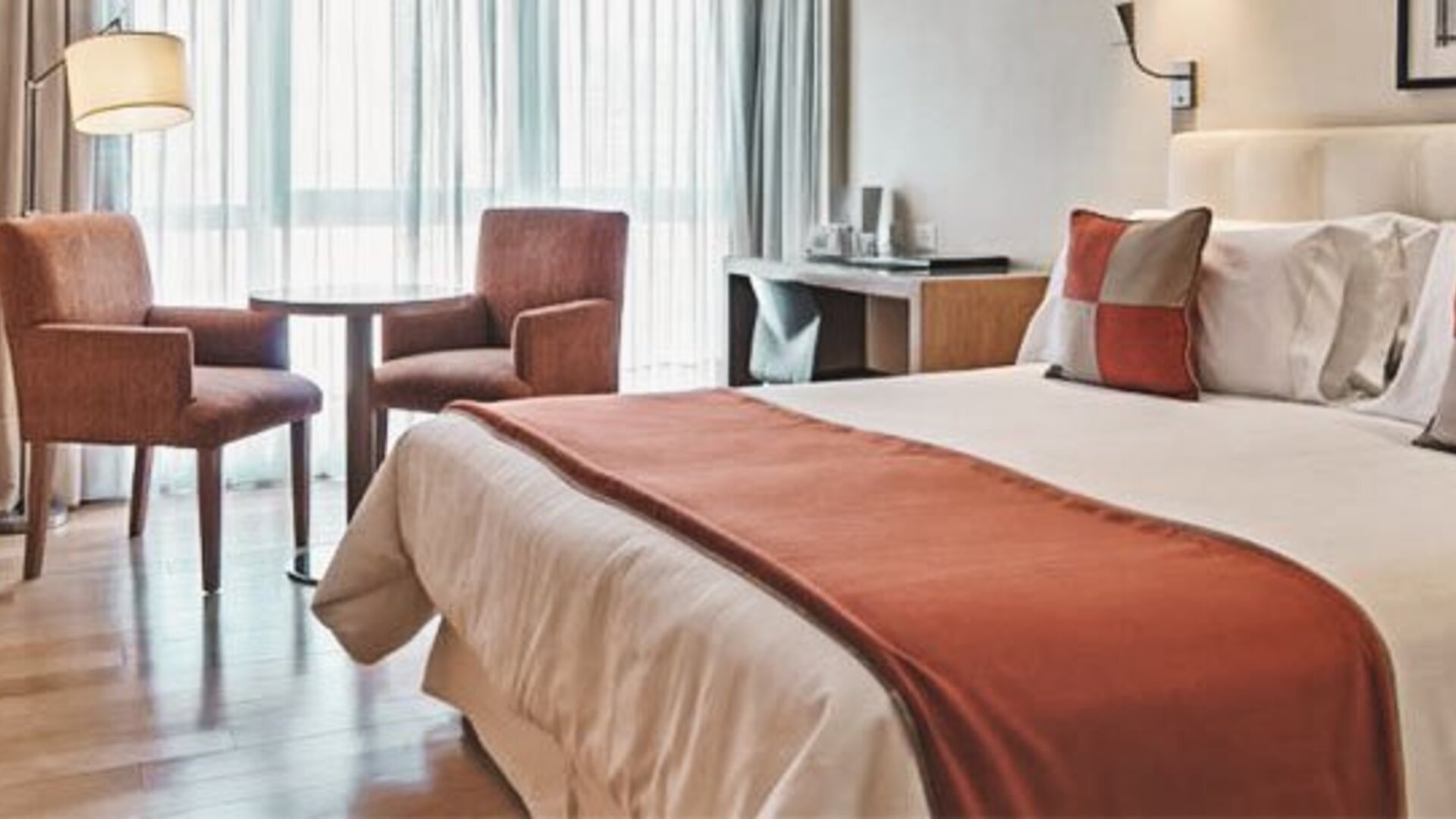 Guest room with double bed, rust-coloured bed runner and cushions, two armchairs and large curtained windows at Hotel Regente Palace.