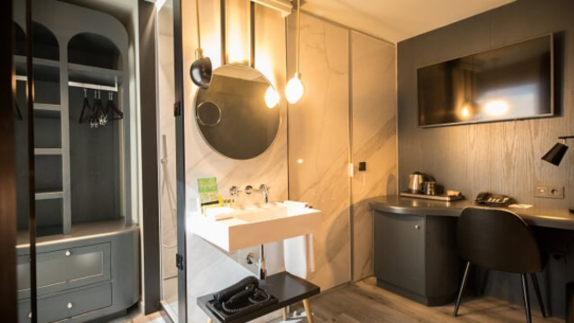 Guest room workspace with wooden desk, chair, wall-mounted television and open wardrobe beside the bathroom at Hotel Silken Saaj Las Palmas.