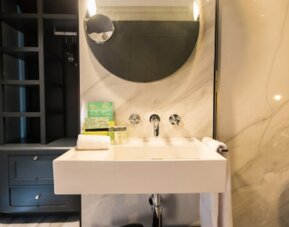 Modern bathroom with white vessel sink, round mirror, marble-effect walls and dark built-in shelving at Hotel Silken Saaj Las Palmas.Meeting room with neatly arranged tables, dark chairs, white place settings and warm recessed lighting at Hotel Silken Saa