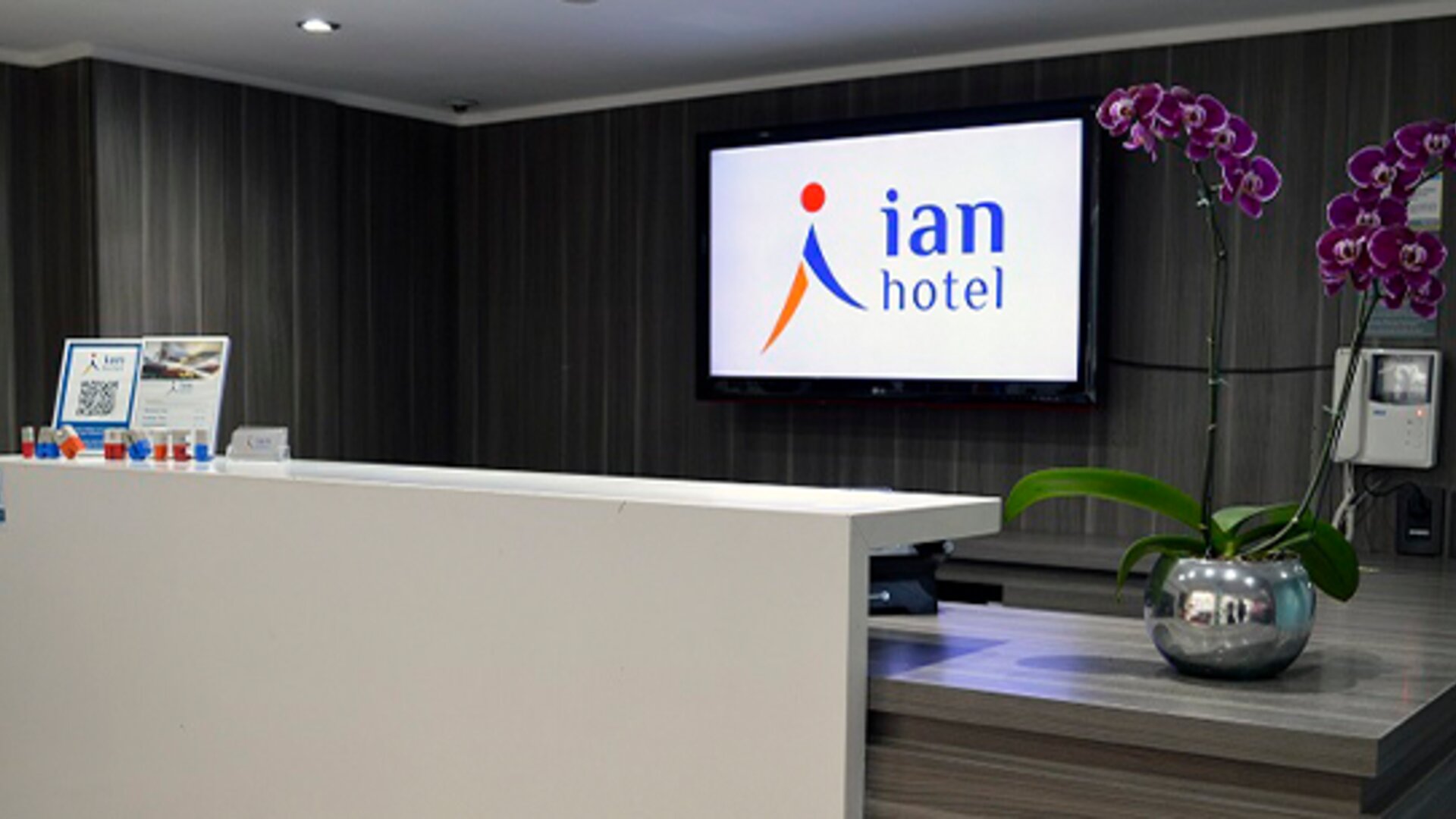 Hotel reception desk with white counter, wall-mounted Ian Hotel logo screen and purple orchids at Ian Hotel.