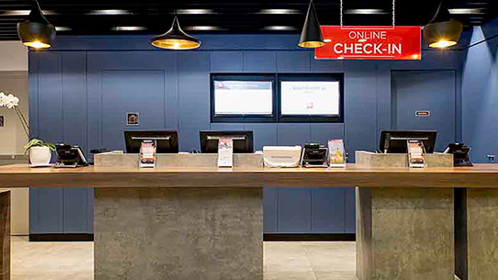 Hotel reception with long wooden counter, blue wall panels, computer stations and hanging pendant lights at Ibis Lima Reducto Miraflores.
