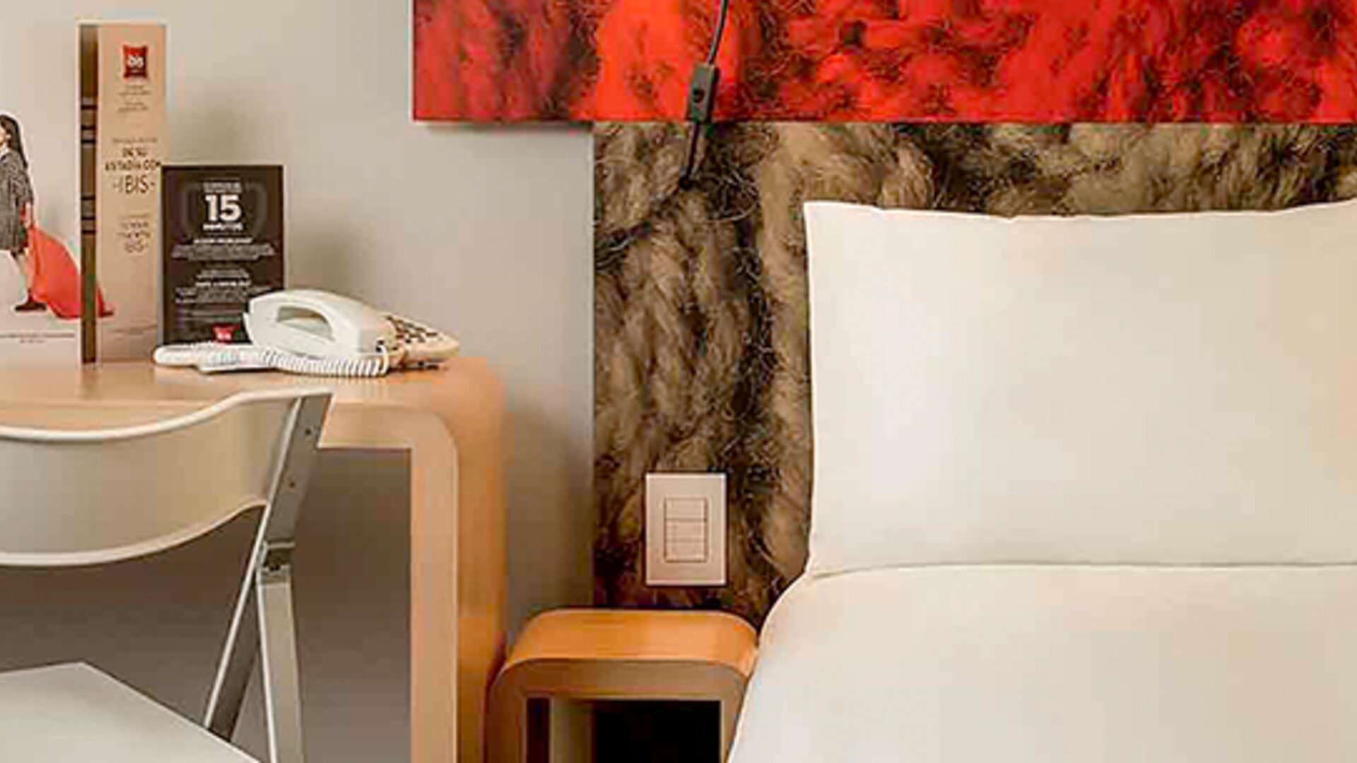 Guest bed with white bedding and pillows beside a wooden desk, chair and bedside table at Ibis Lima Reducto Miraflores.
