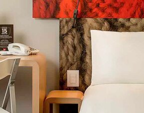 Guest bed with white bedding and pillows beside a wooden desk, chair and bedside table at Ibis Lima Reducto Miraflores.