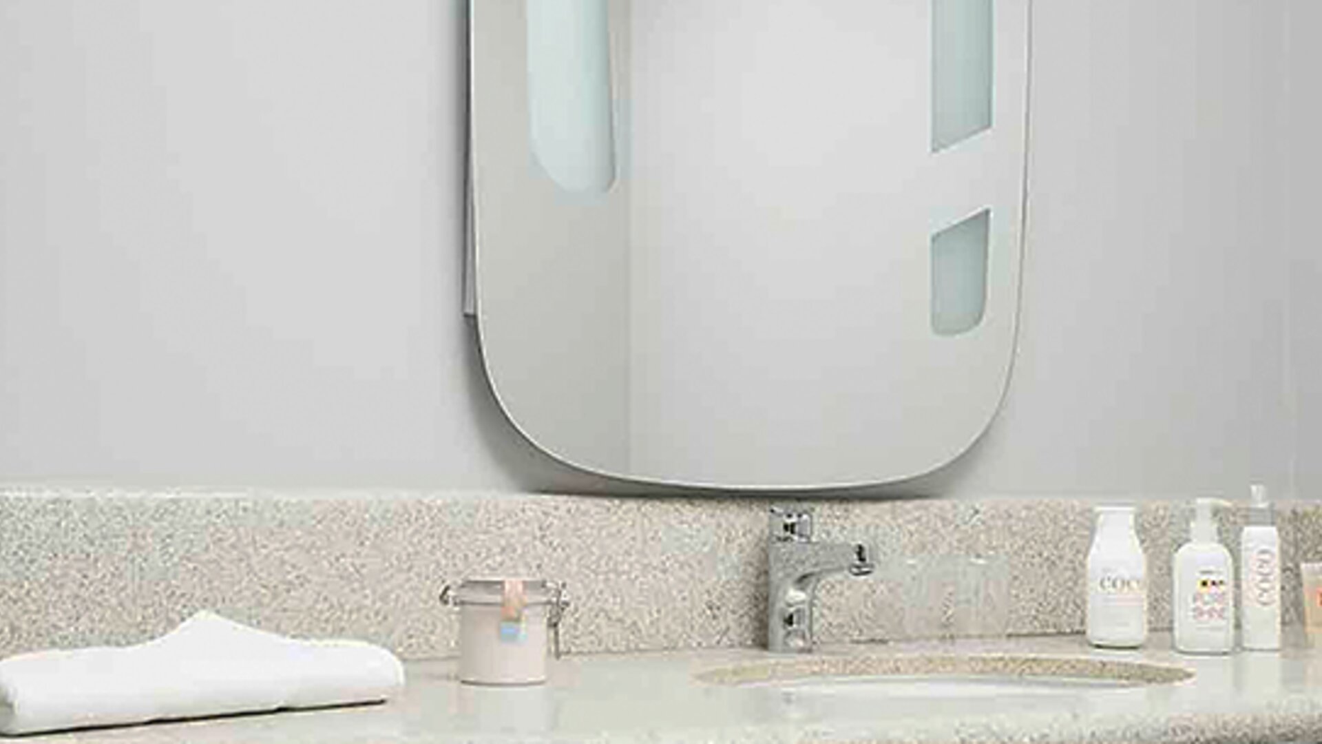 Bathroom vanity with speckled countertop, white sink, wall mirror, toiletries and folded towel at Ibis Lima Reducto Miraflores.
